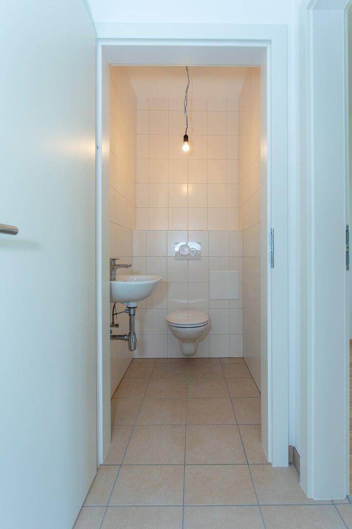 Kleines Gäste-WC mit weißen Wandfliesen, beigefarbenem Boden, Toilette und einem Waschbecken.