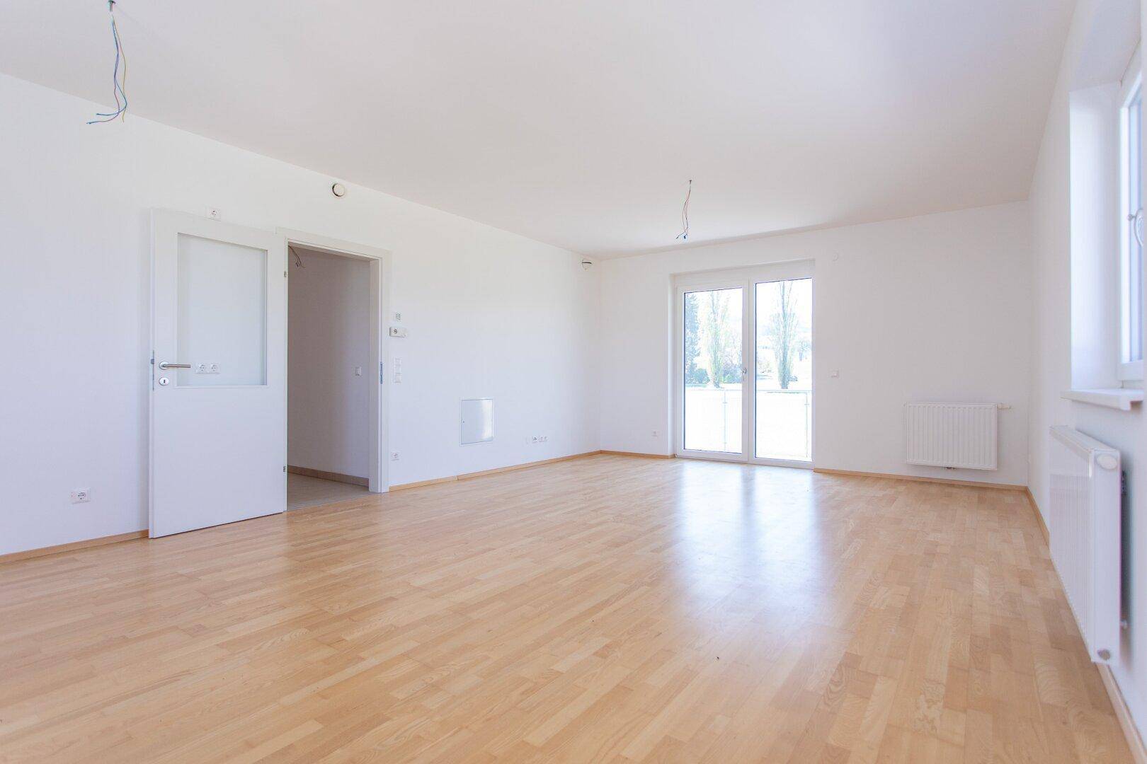 Helles, leeres Zimmer mit hellem Parkettboden, weißen Wänden und Zugang zu einem Balkon.