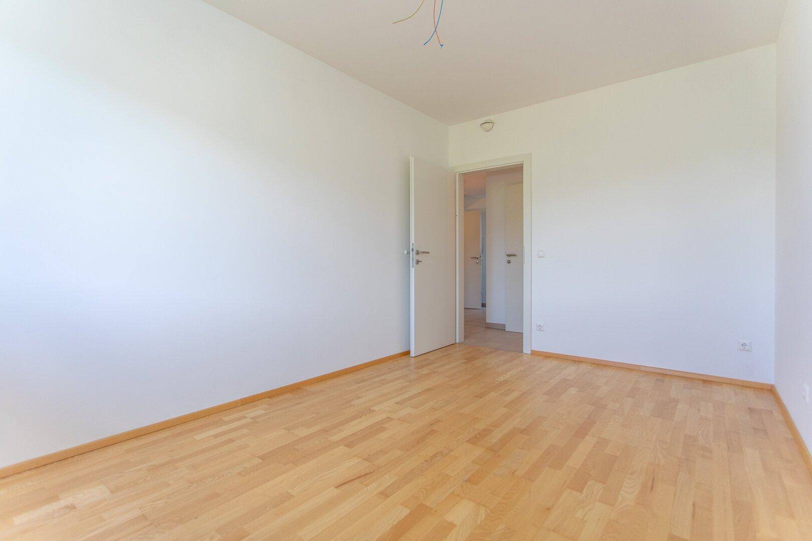 Leeres Schlafzimmer mit Parkettboden und weiß gestrichenen Wänden, mit Zugang zu einem Flur.