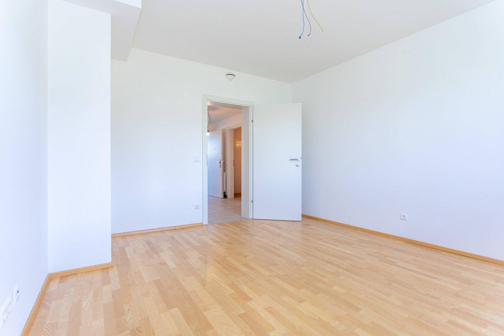 Geräumiges Schlafzimmer mit Parkettboden und weißen Wänden, mit offenem Zugang zum Flurbereich.