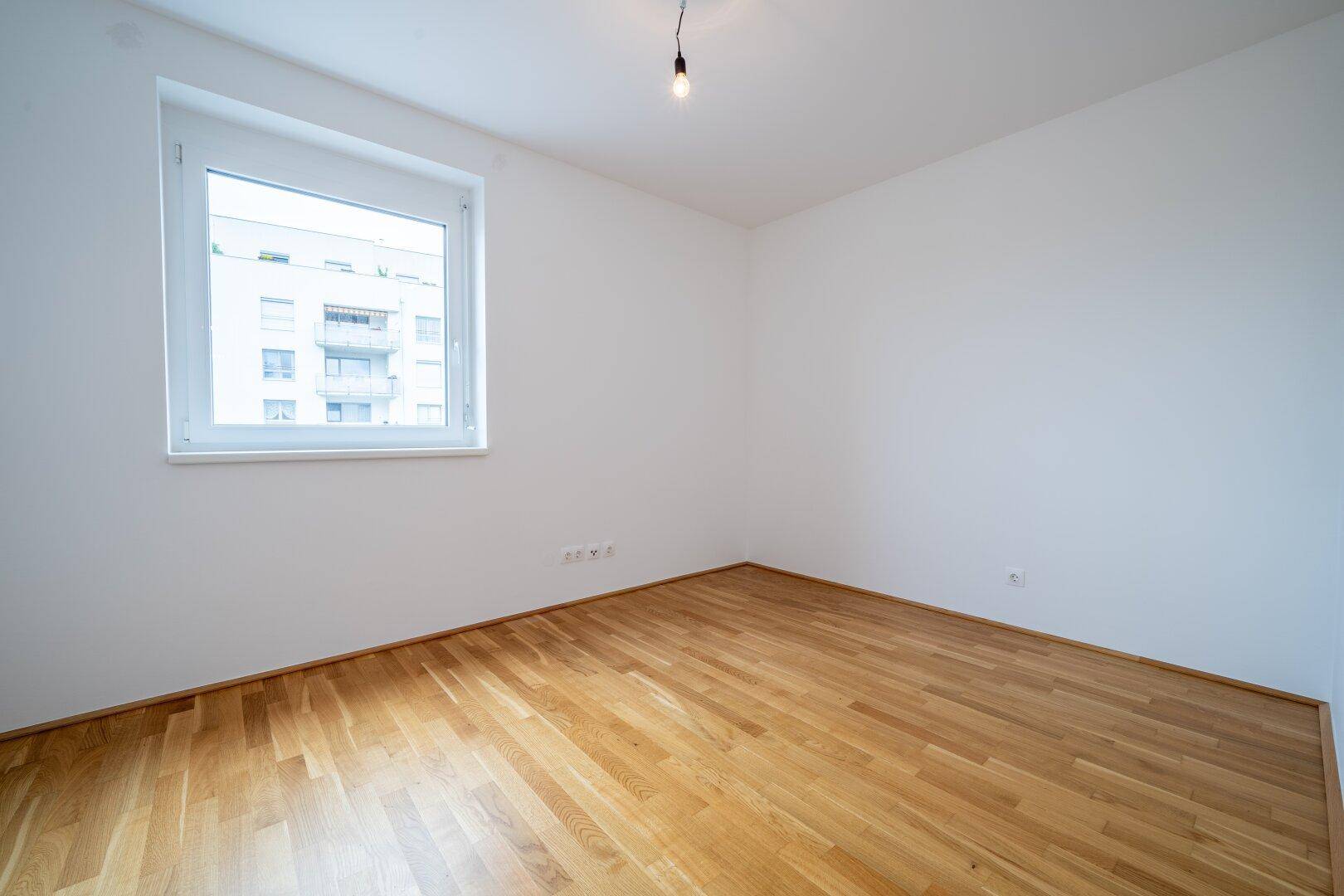 Helles Schlafzimmer mit Parkettboden und einem Fenster, das auf die umliegenden Gebäude blickt.