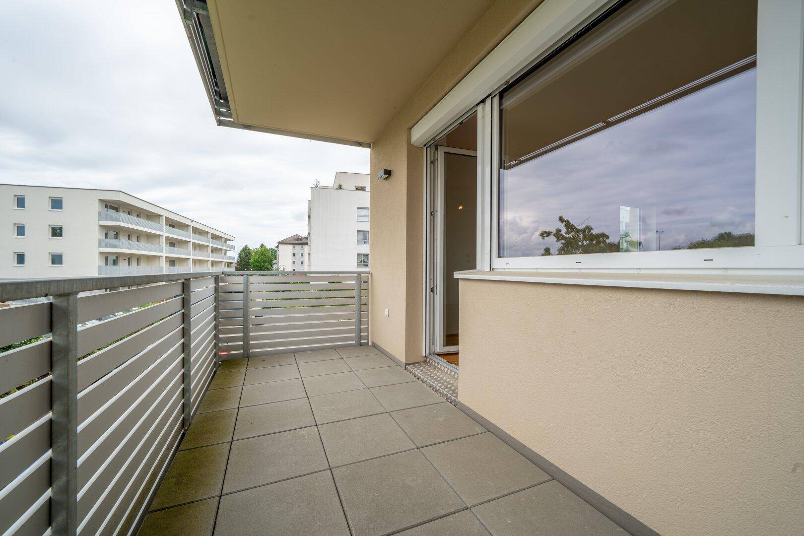 Moderner Balkon mit Fliesenboden und Rollladen am Fenster, mit Blick auf andere Gebäude.