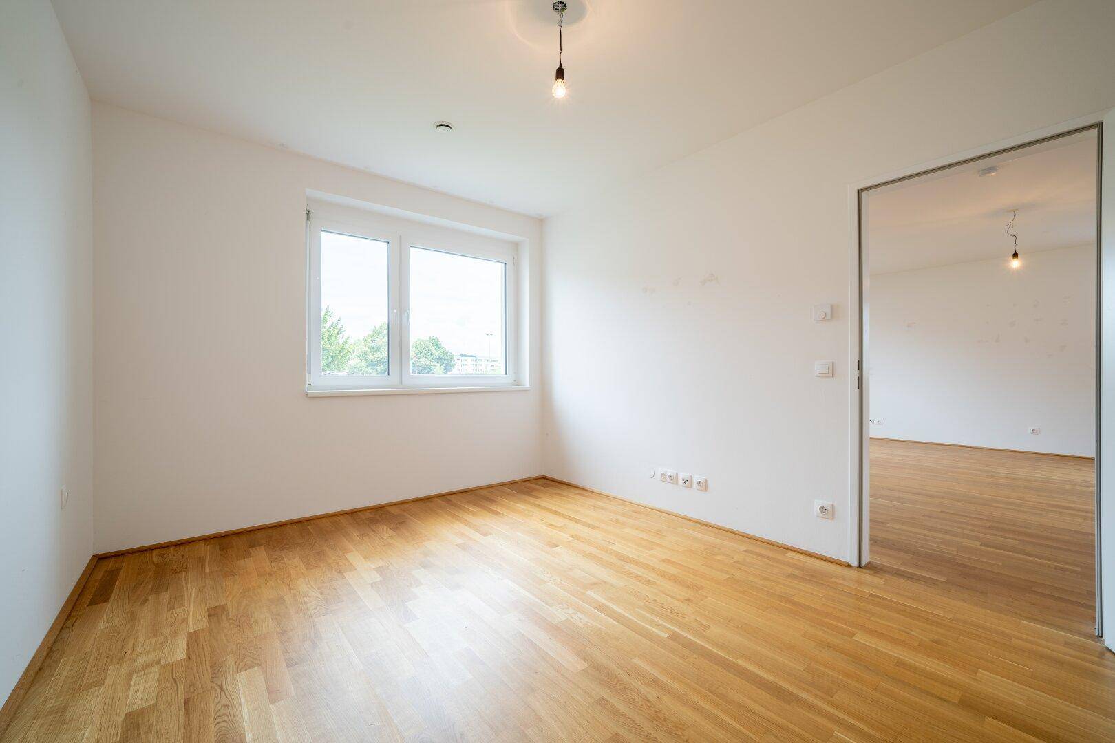 Leeres Zimmer mit Parkettboden und einem Fenster, das eine angenehme Helligkeit bietet.
