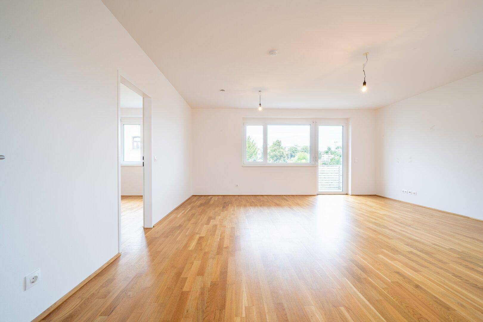 Geräumiges Wohnzimmer mit hellem Parkettboden und großen Fenstern, die viel Tageslicht hereinlassen.