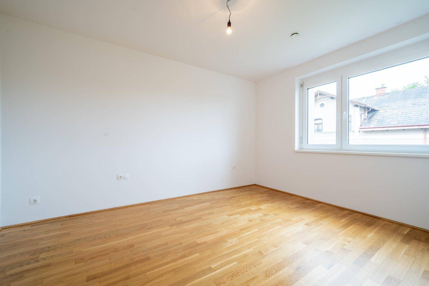 Leeres Zimmer mit Parkettboden und einem Fenster, das den Blick nach draußen freigibt.