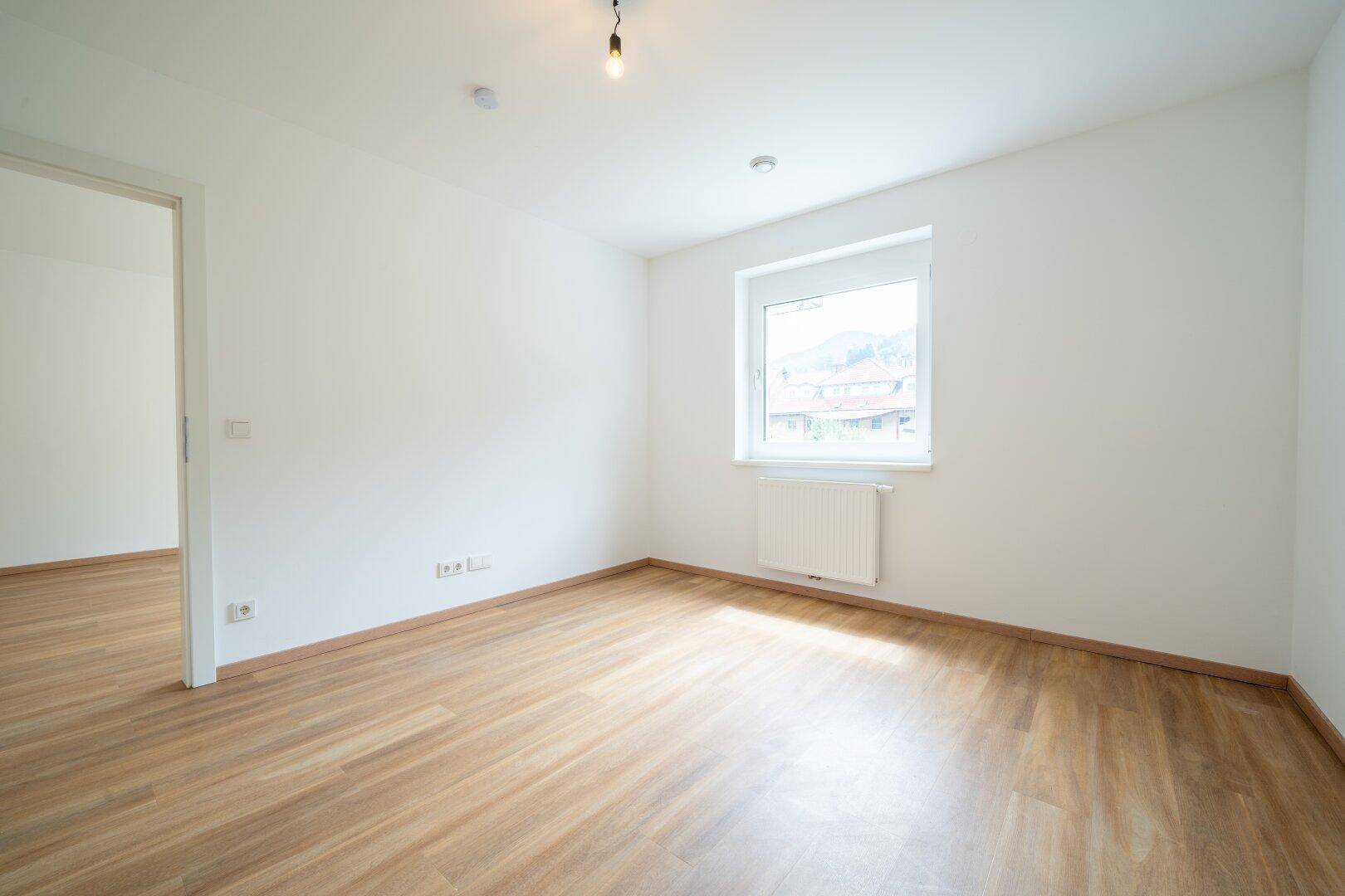 Helles Schlafzimmer mit Holzboden, weiß gestrichenen Wänden und einem Fenster mit Ausblick.