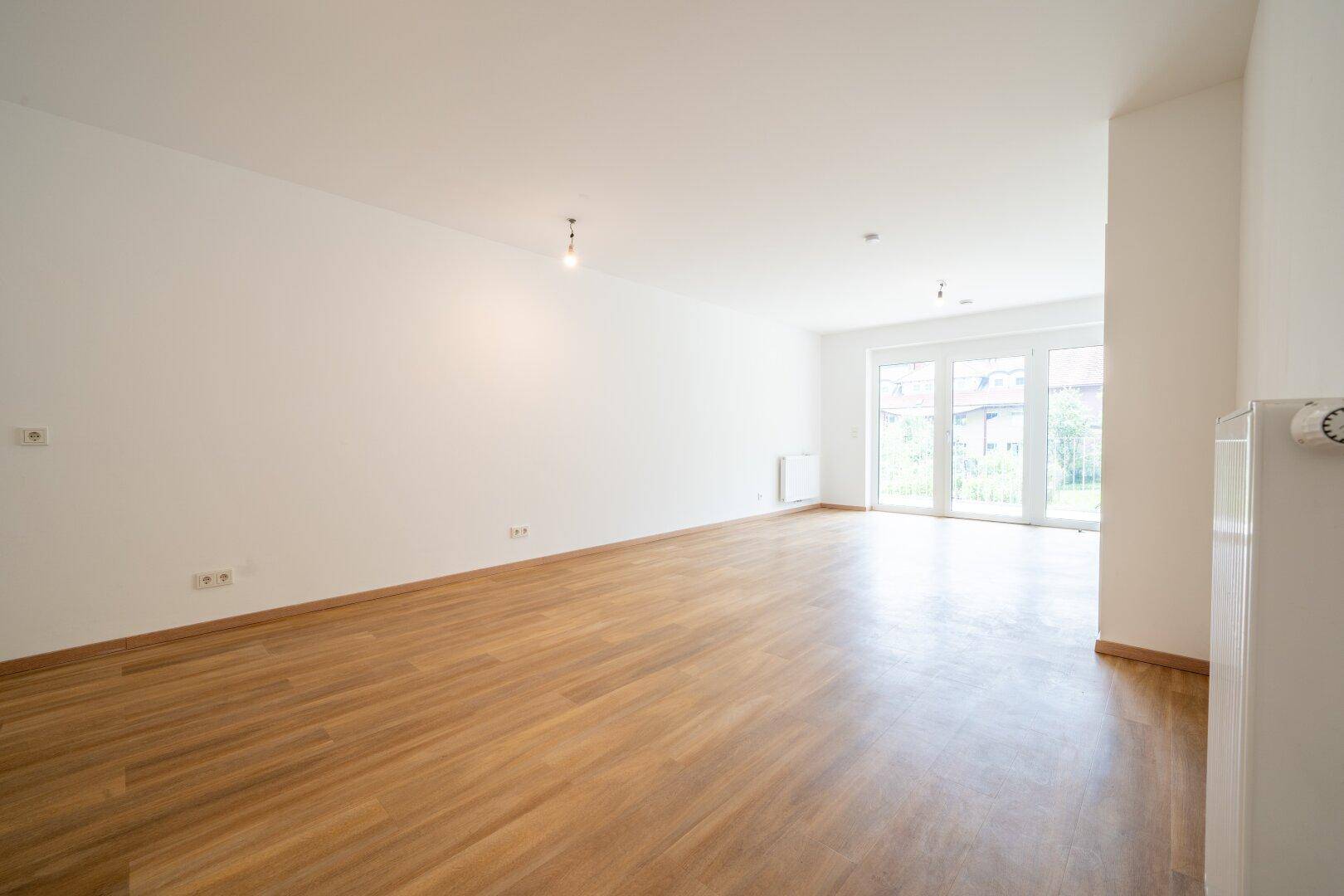 Helles, leeres Wohnzimmer mit modernem Holzboden und Zugang zu einem Außenbereich.