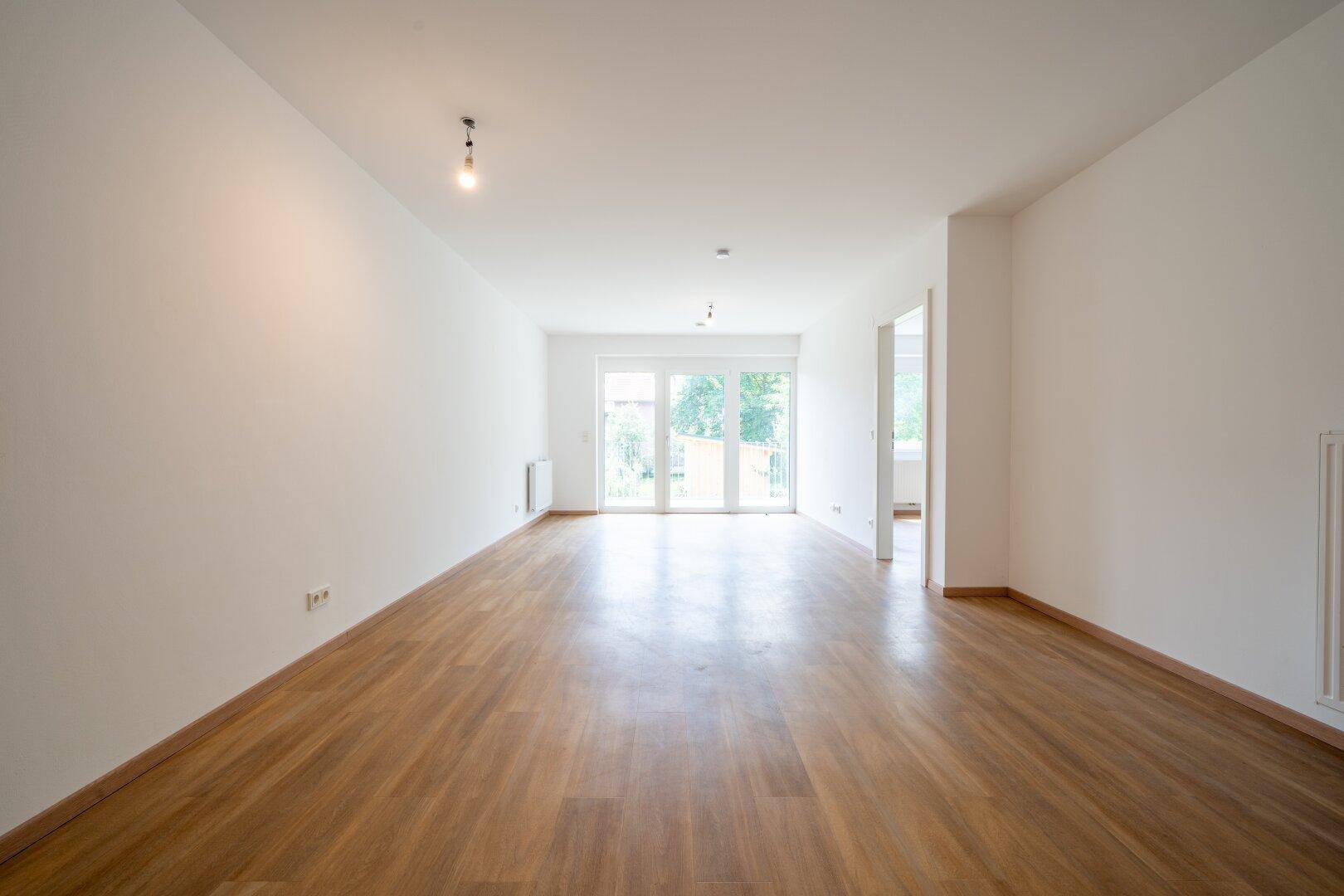 Großzügiges, helles Wohnzimmer mit Holzboden und großen Fenstertüren, die viel Licht hereinlassen.