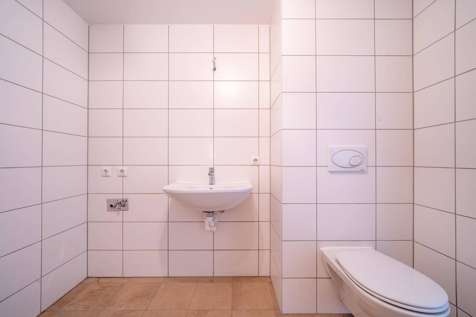 Helles Badezimmer mit weißen Fliesen, wandhängendem WC und Waschbecken, in gutem Zustand.