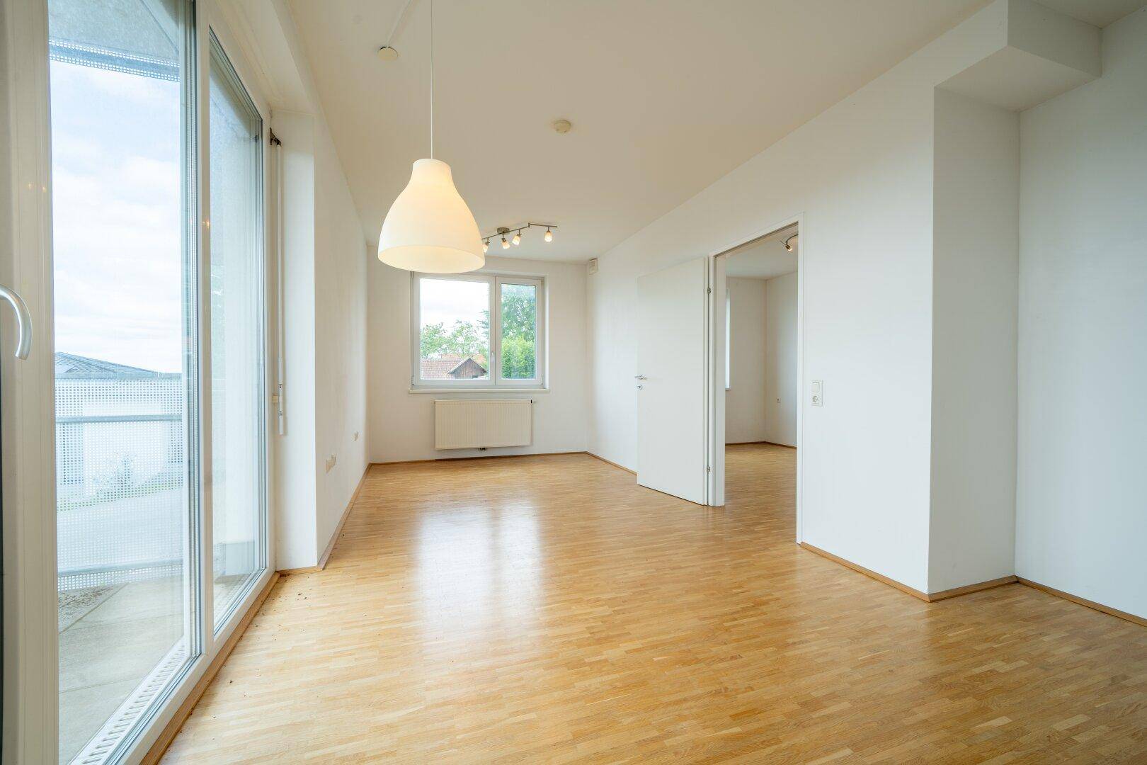 Heller Wohnbereich mit Parkettboden und Zugang zum Balkon, bietet Ausblick ins Grüne.