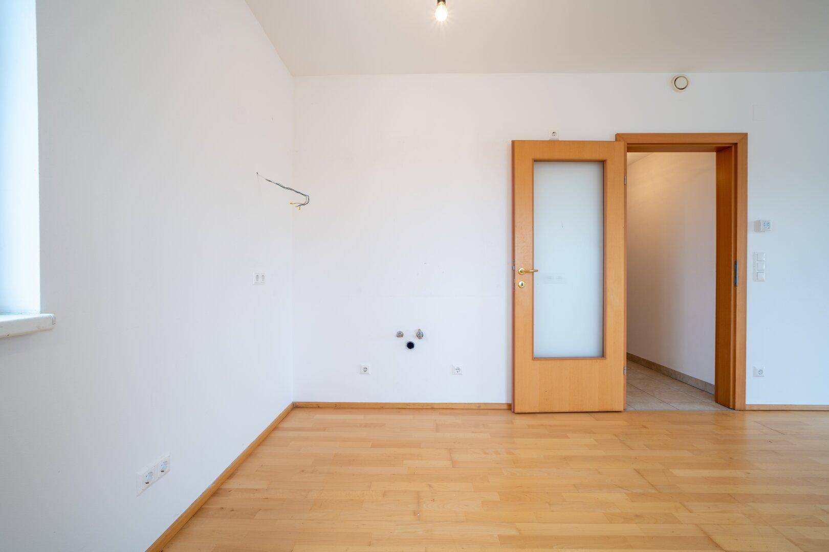 Leerer Raum mit Parkettboden und Anschlüssen an der Wand, möglicherweise für eine Küche oder Waschmaschine.