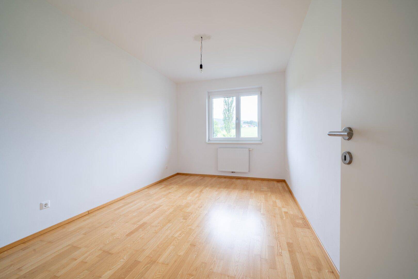 Ruhiges Schlafzimmer mit hellem Parkettboden und einem Fenster, das einen Blick ins Grüne bietet.