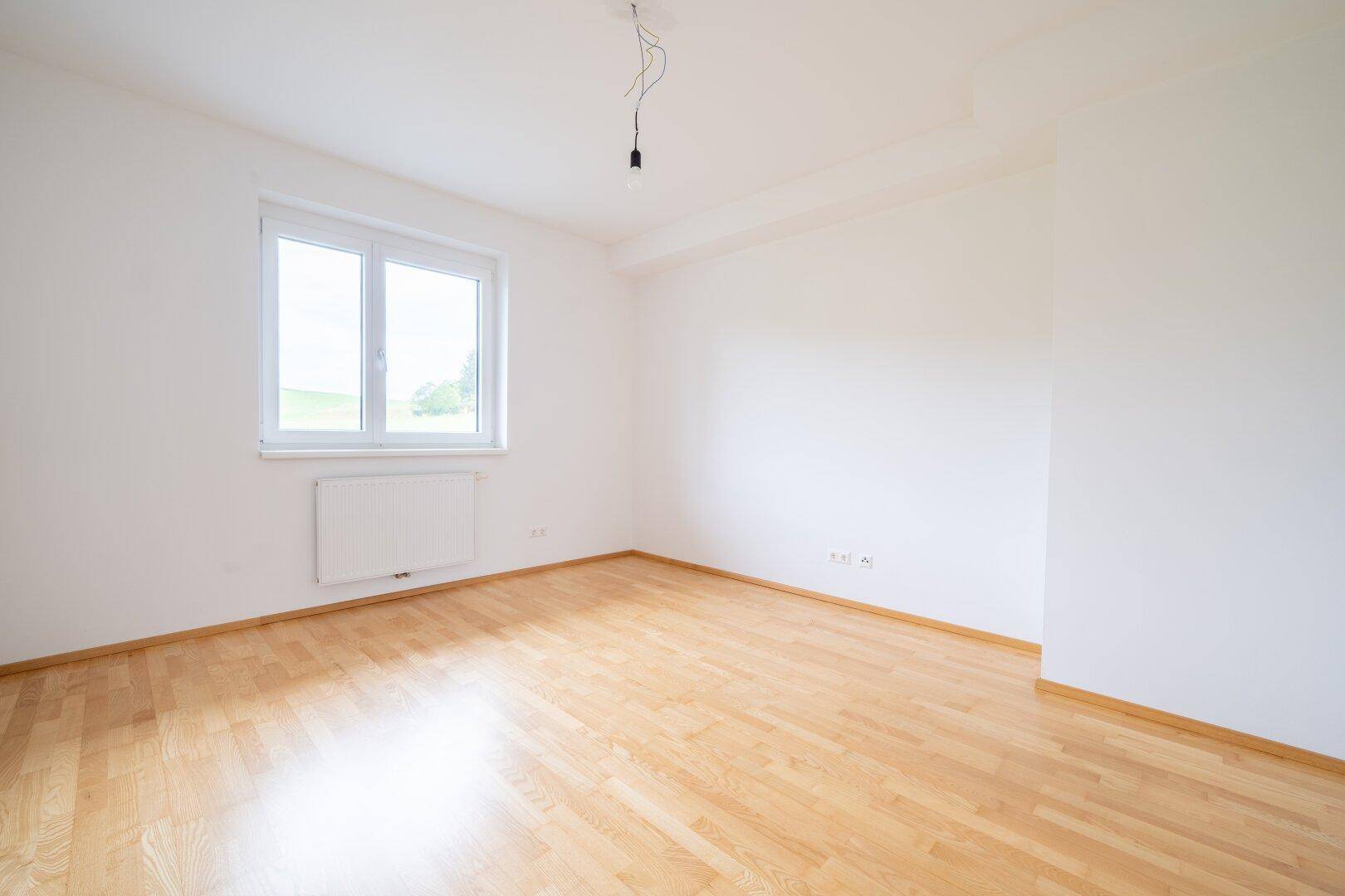 Helles Schlafzimmer mit Parkettboden und einem Fenster, das einen weiten Blick in die Landschaft ermöglicht.