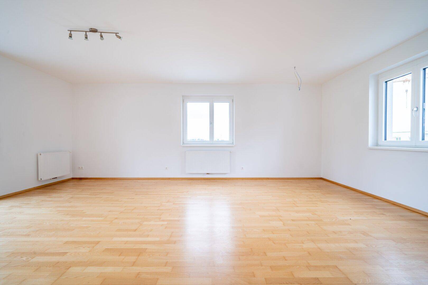 Geräumiges Wohnzimmer mit hellem Parkettboden und zwei Fenstern, die viel Tageslicht hereinlassen.