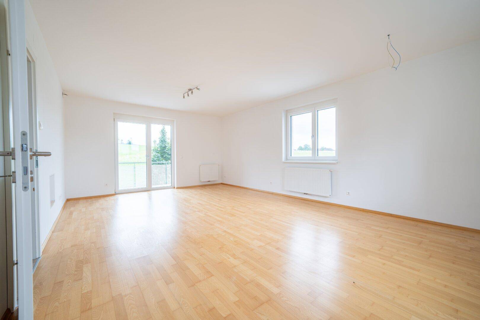 Helles Wohnzimmer mit Parkettboden, weißen Wänden und Zugang zum Balkon mit grüner Aussicht.