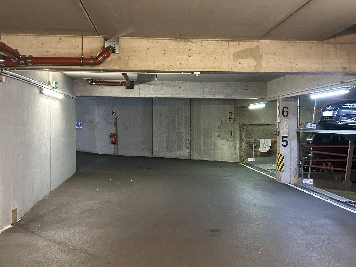 Weitläufiger Bereich einer Tiefgarage mit Betonwänden, sichtbaren Rohren und einer Rampe.