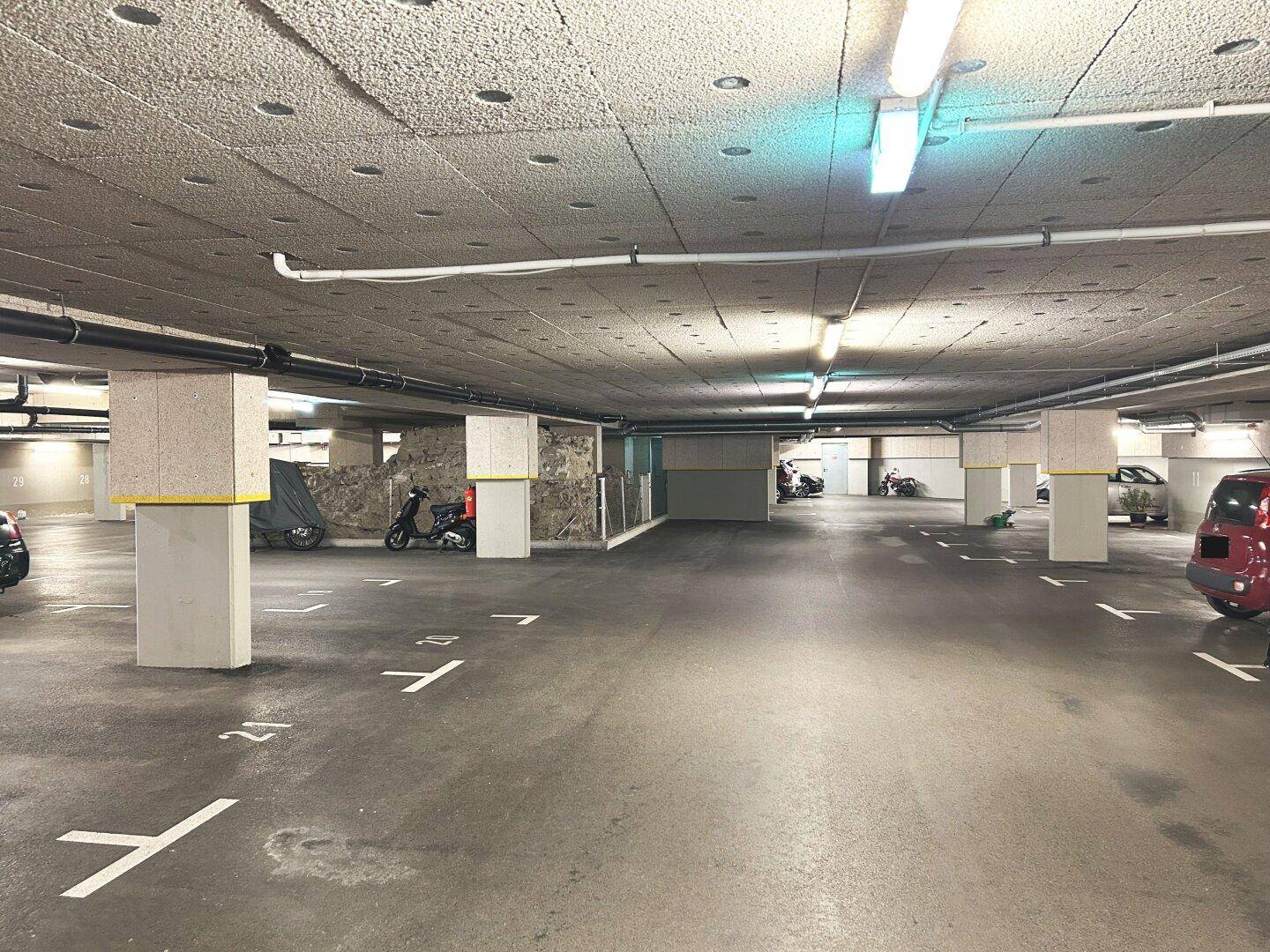 Geräumige Tiefgarage mit zahlreichen Stellplätzen, Betonpfeilern und sichtbaren Installationen an der Decke.