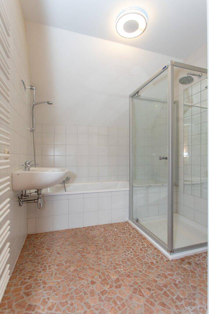 Helles Badezimmer mit Badewanne, Duschkabine und Heizkörper, ausgestattet mit weißen Fliesen und Mosaikboden.