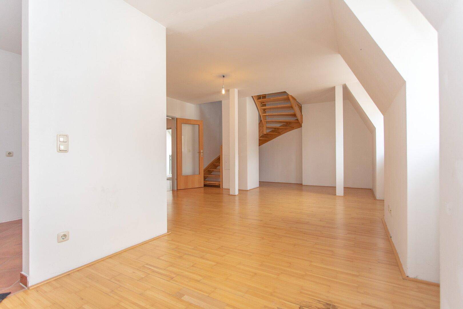 Heller Wohnraum mit Parkettboden, Dachschrägen und einer geschwungenen Holztreppe, die den oberen Bereich erschließt.