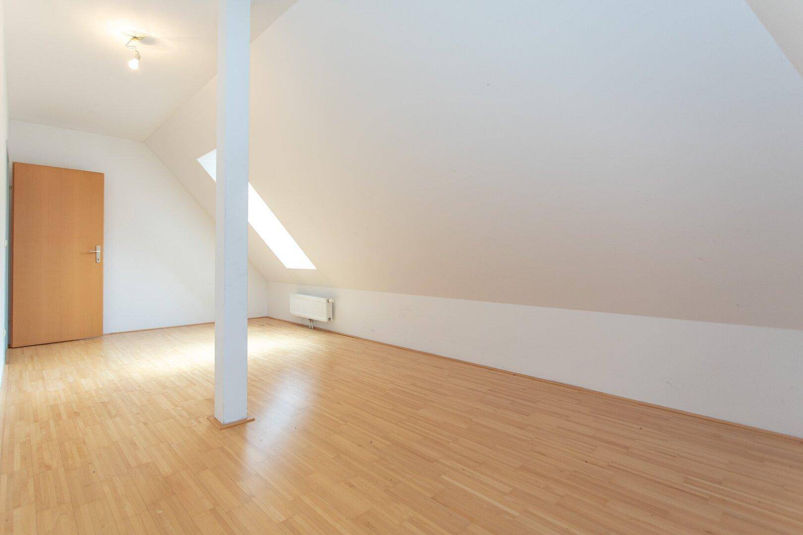Geräumiges Dachzimmer mit Parkettboden, Dachschrägen und einem kleinen Dachfenster, das den Raum erhellt.