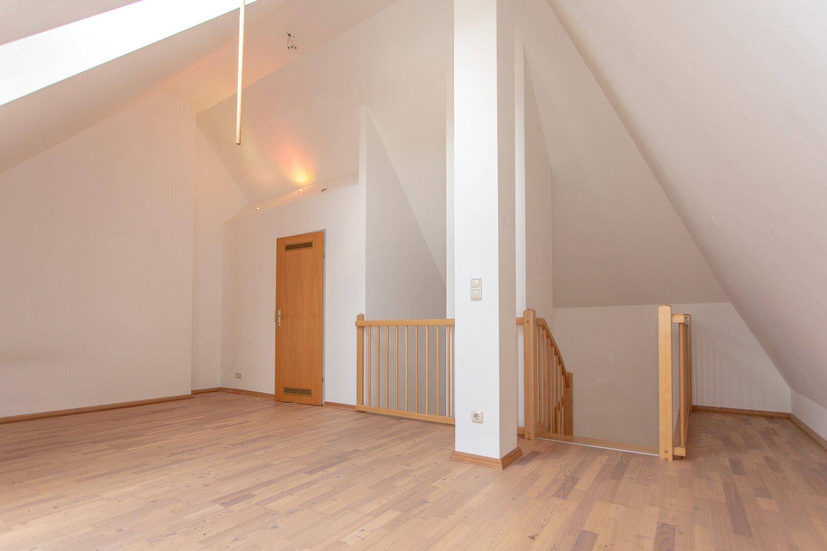 Helles Dachzimmer mit Parkettboden, Dachschrägen und einem Treppengeländer, das den Zugang sichert.