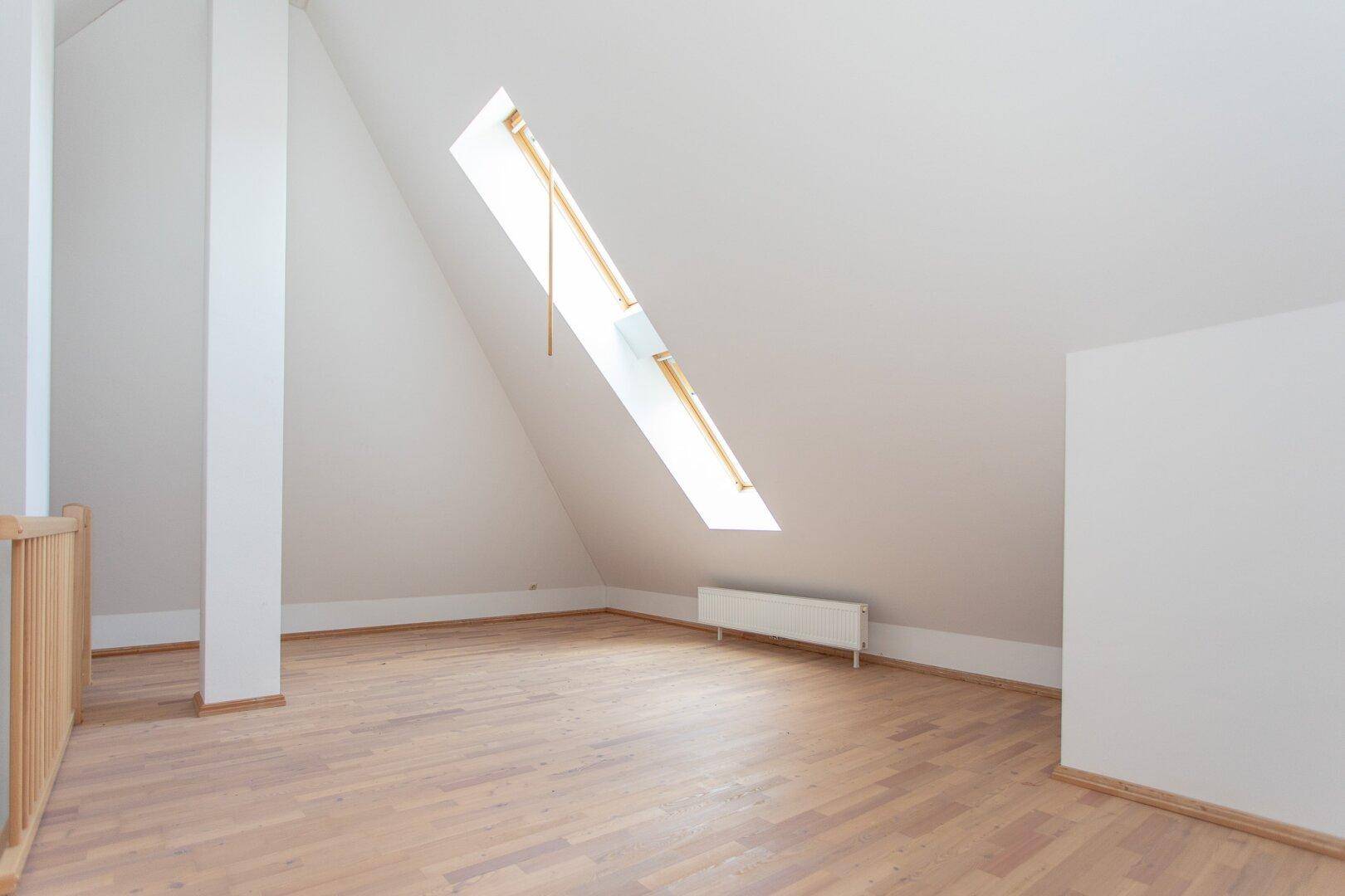 Dachzimmer mit Parkettboden, Dachschräge und einem großen Dachfenster, das den Raum mit Licht füllt.