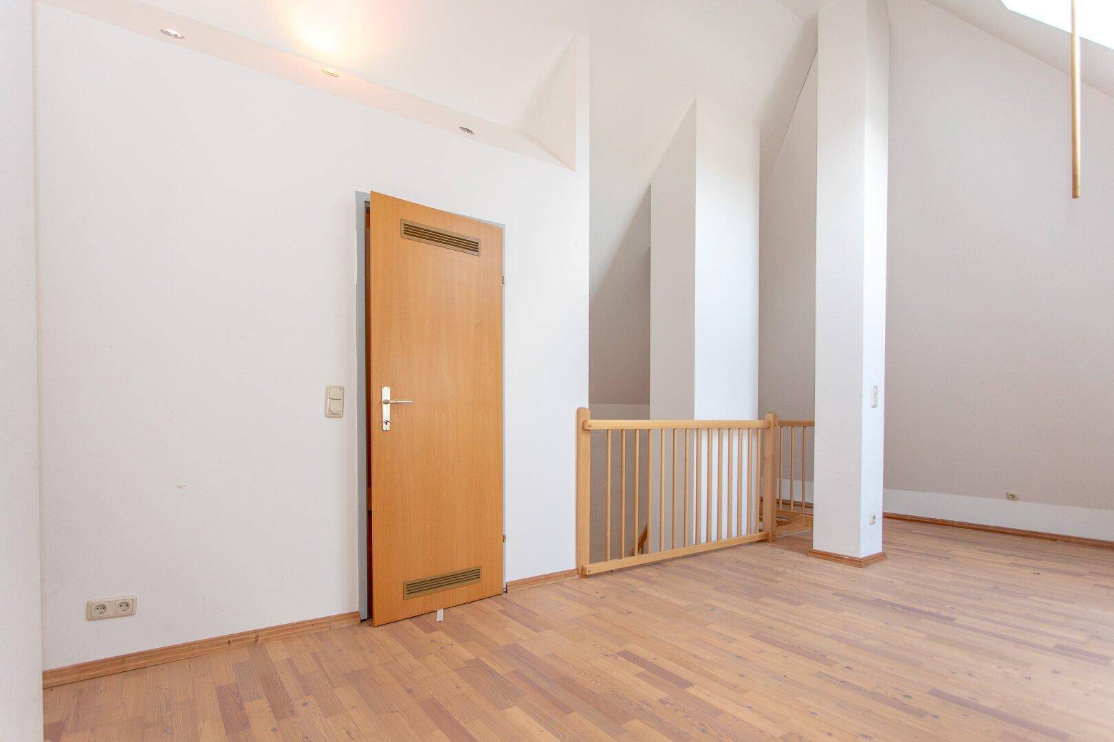 Geräumiges Dachzimmer mit Parkettboden, Dachschrägen und einer Holztür, die zu einem weiteren Raum führt.