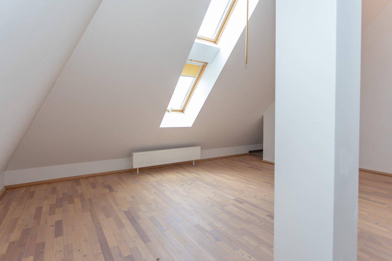 Helles Dachzimmer mit Parkettboden, Dachschrägen und zwei Dachfenstern, die für optimale Belichtung sorgen.