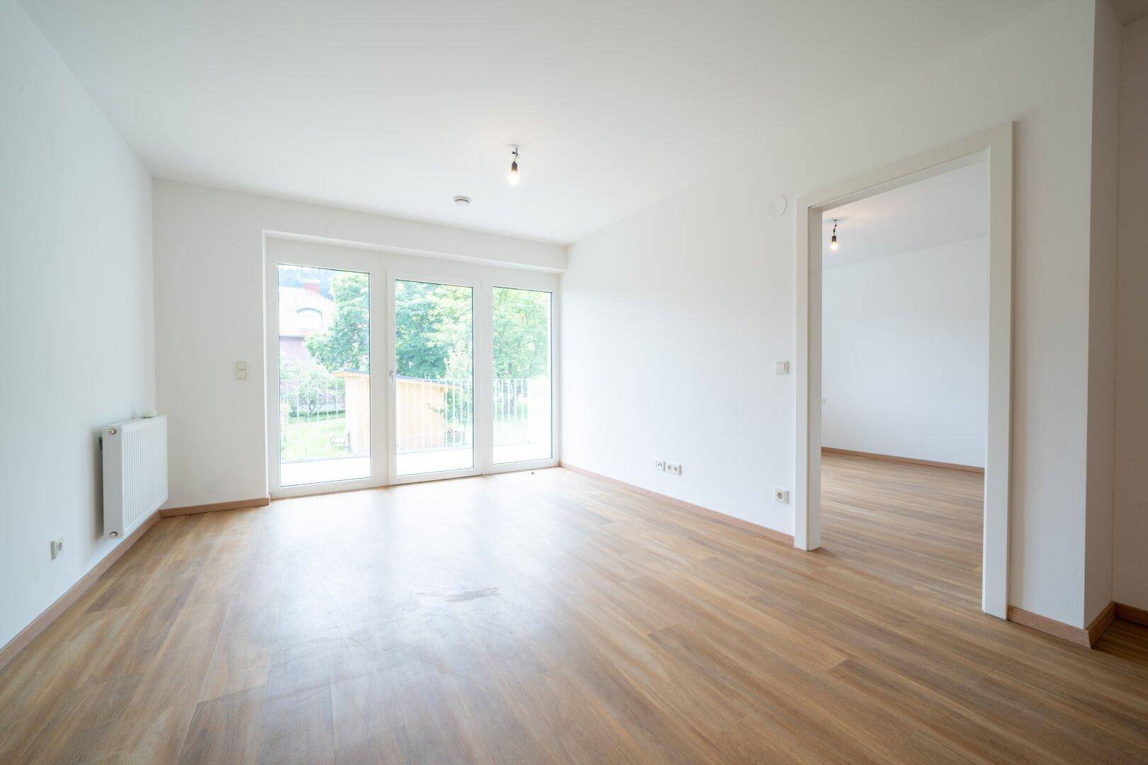 Lichtdurchfluteter Raum mit Laminatboden und großen Fenstern, die den Blick ins Grüne freigeben.
