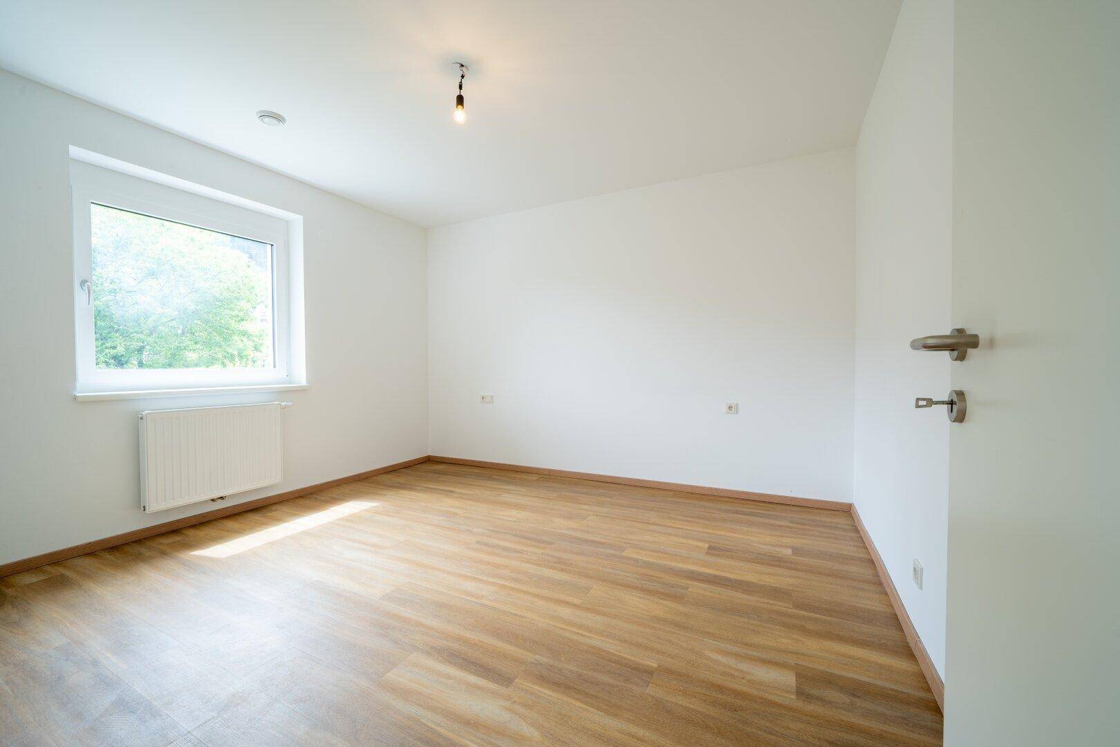 Ruhiges Schlafzimmer mit Laminatboden und einem Fenster mit Blick ins Grüne.