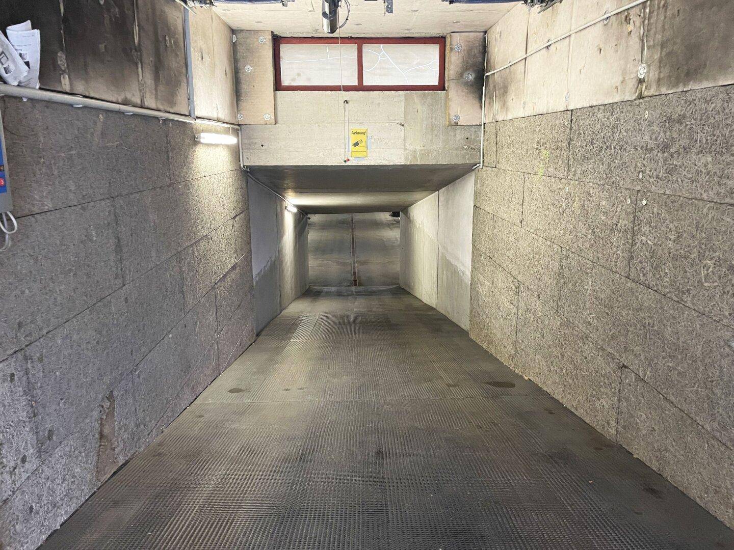 Langer, schmaler Tiefgaragentunnel mit Betonsteinwänden und Metallgitterboden, gut beleuchtet.