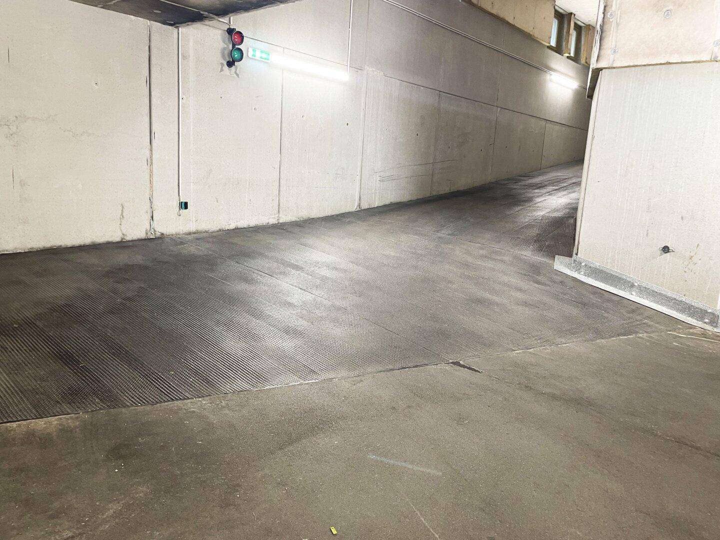 Abfallende Tiefgaragenrampe mit Betonwänden und strukturierter Fahrbahn, beleuchtet von Leuchten.