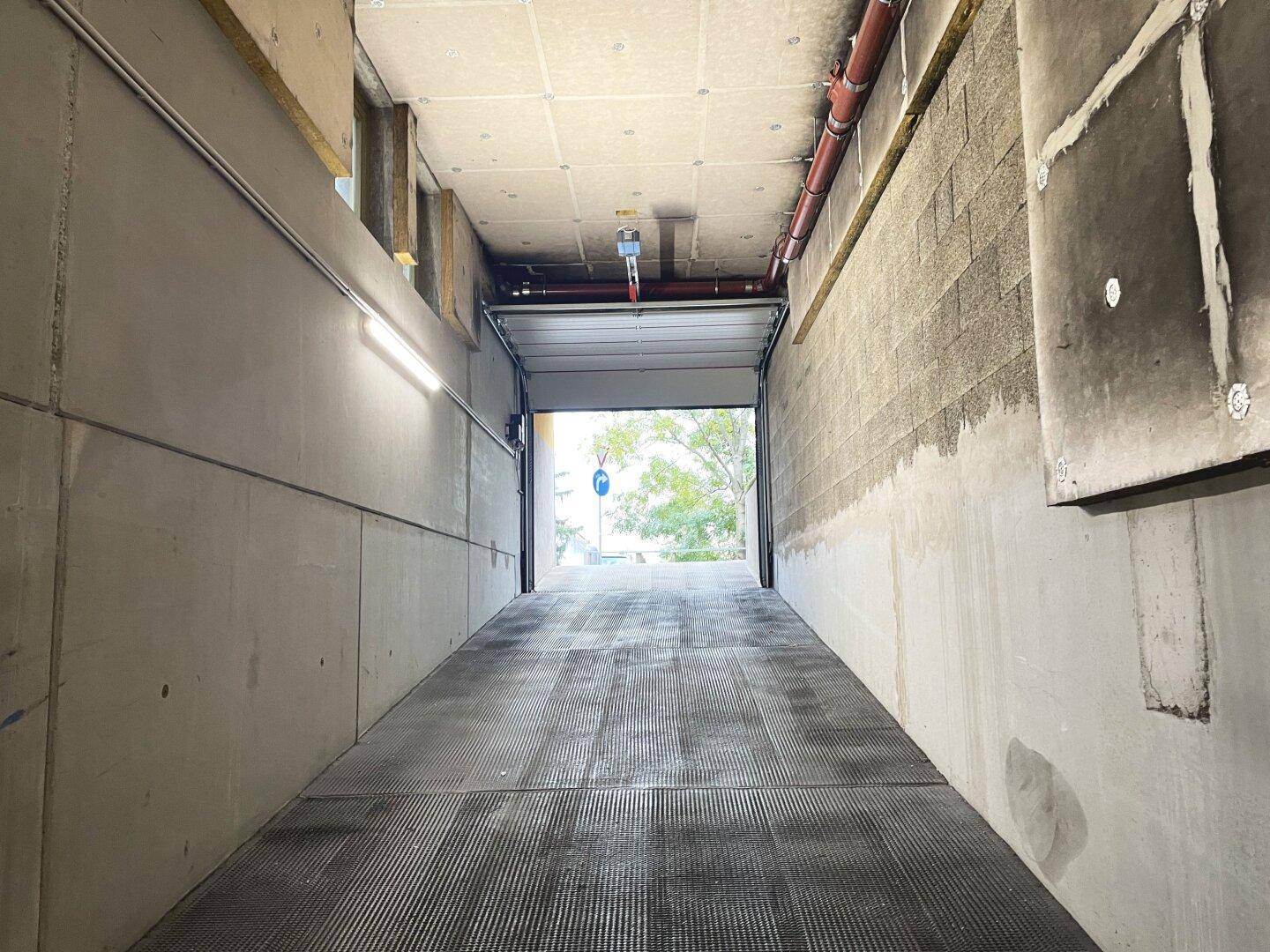 Einfahrt zu einer Tiefgarage mit Betonwänden und Metallgitterboden, die ins Freie führt.