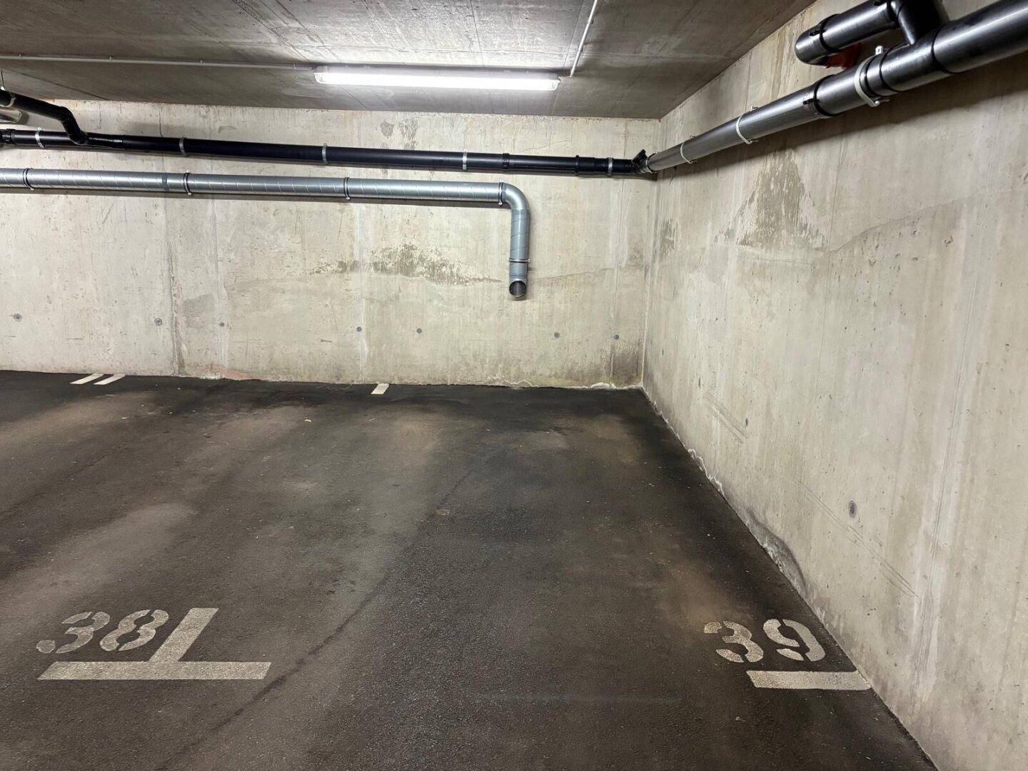 Nahaufnahme der Betonwand und der sichtbaren Versorgungsleitungen in der Tiefgarage mit Stellplatzmarkierungen.