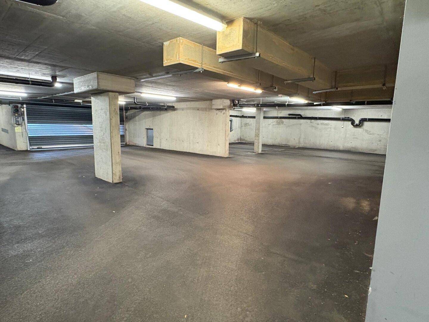 Geräumige Tiefgarage mit Betonpfeilern, Lüftungskanälen und einem Rolltor am Ende des Raumes.