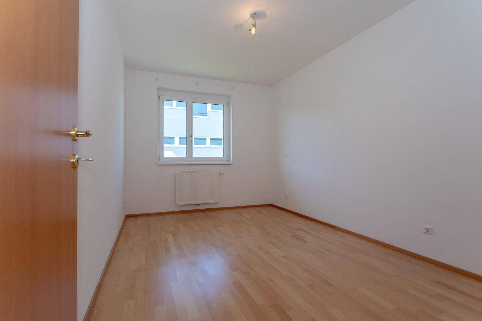 Helles Schlafzimmer mit Parkettboden und einem Fenster mit Blick auf ein Nachbargebäude.