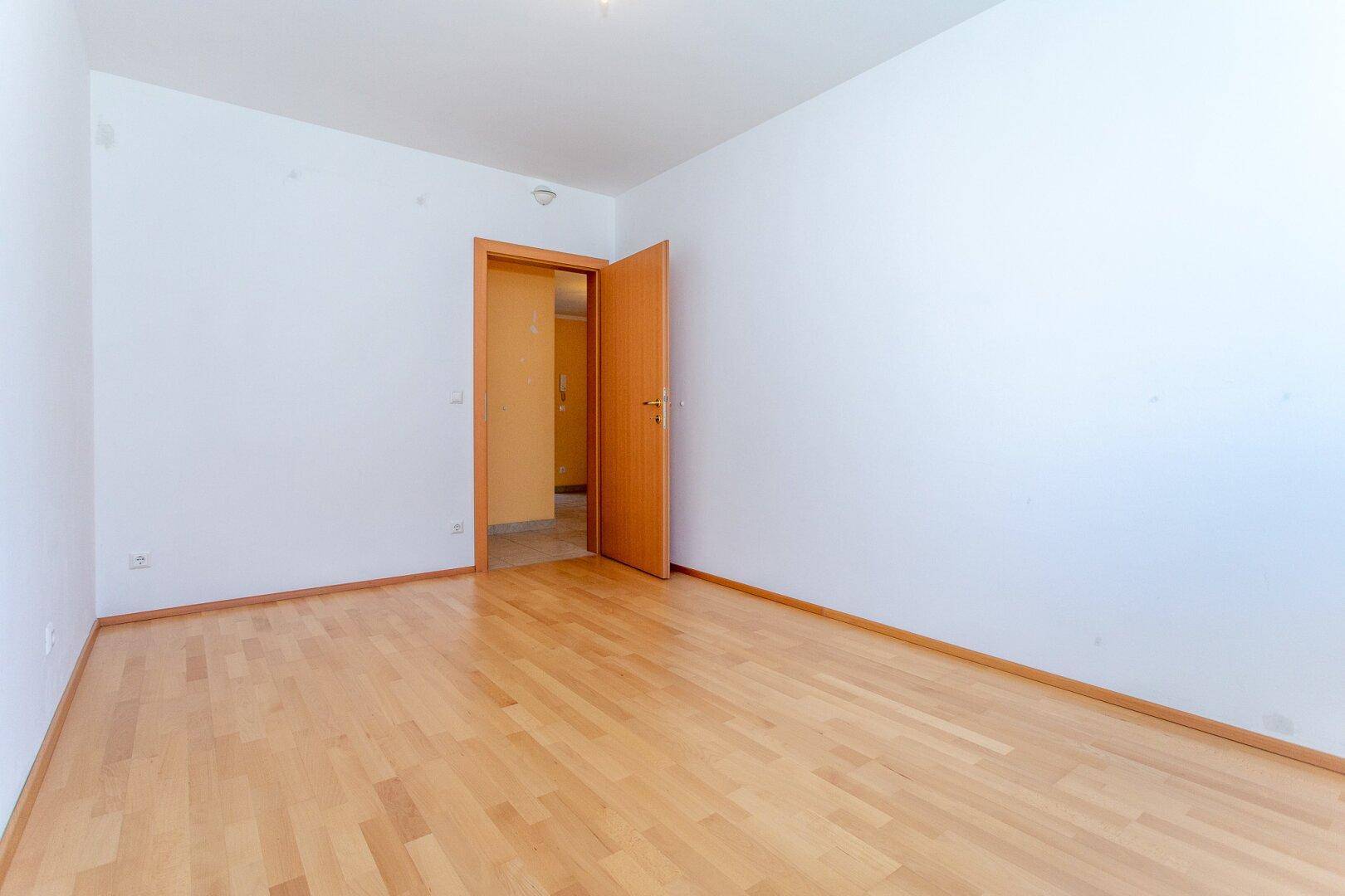 Leeres Zimmer mit Parkettboden und einer offenen Tür, die in einen Flur führt.
