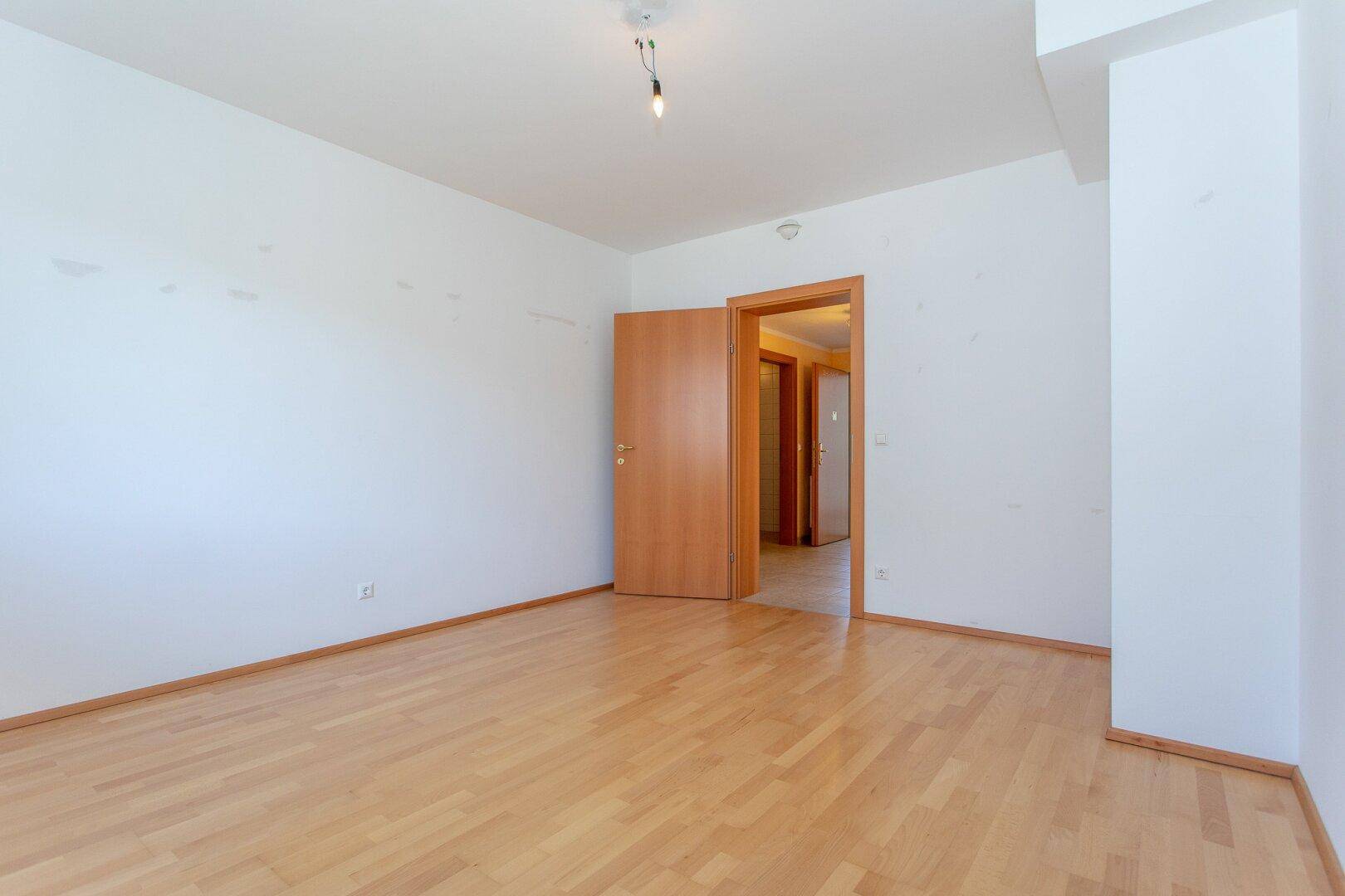 Helles Zimmer mit Parkettboden und offener Tür zu einem weiteren Raum.