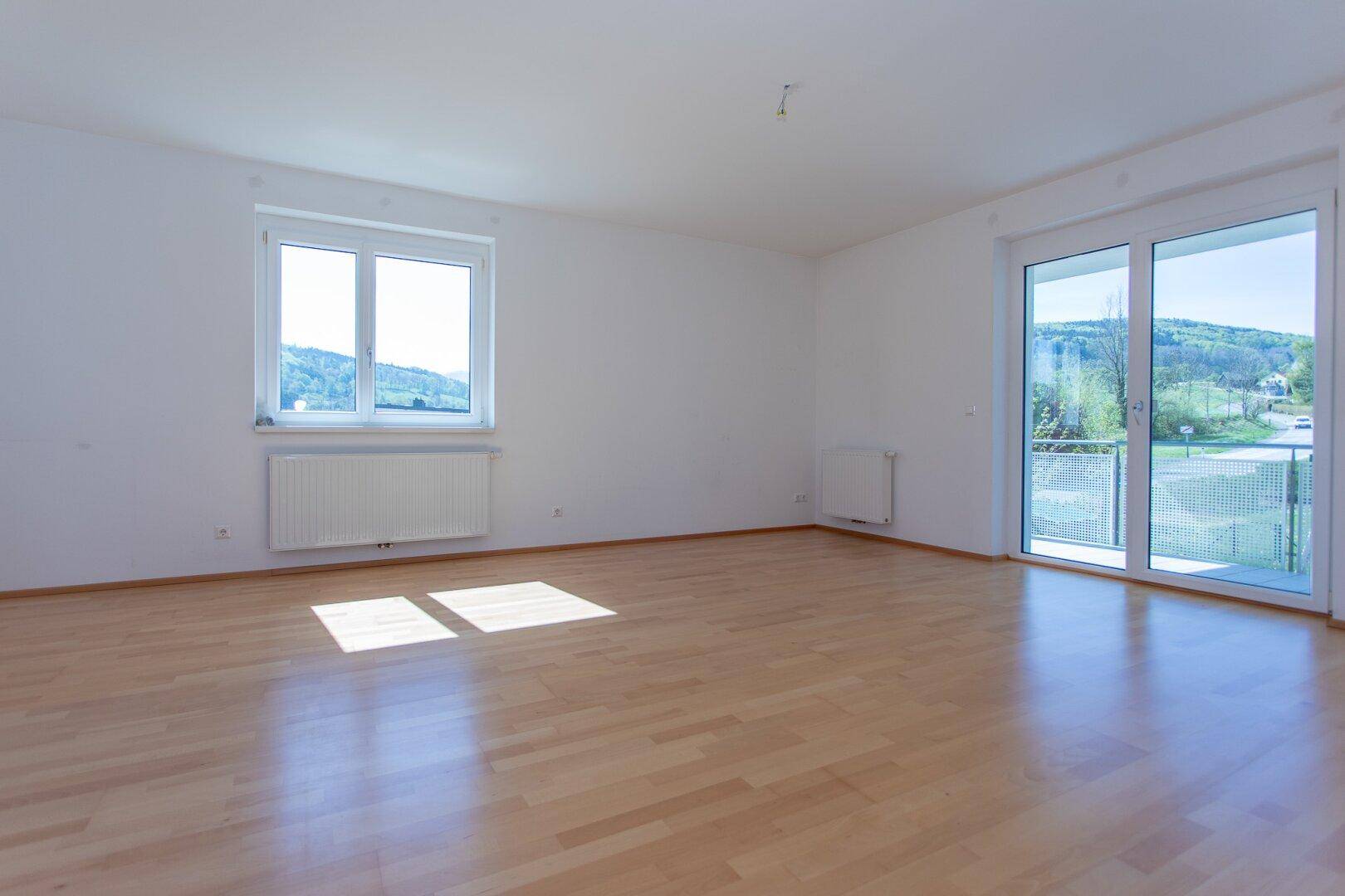 Helles, leeres Wohnzimmer mit Parkettboden und Zugang zu einem Balkon mit Weitblick.