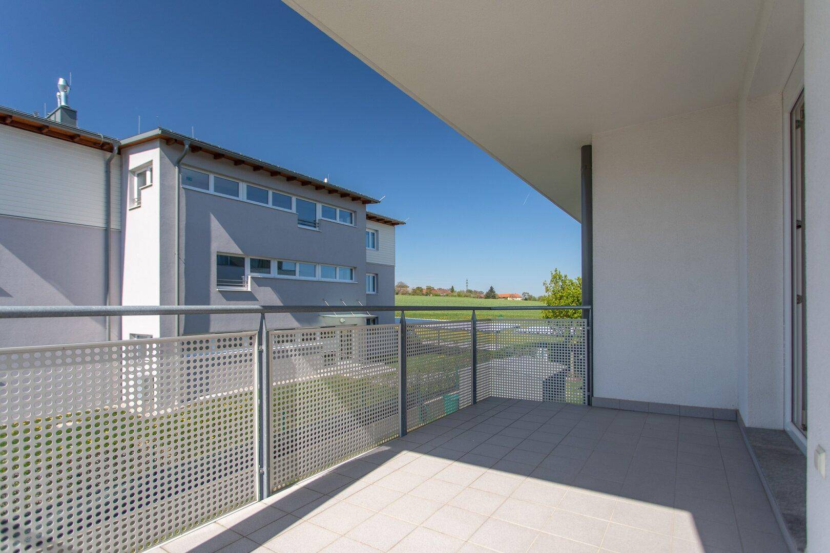 Balkon mit Fliesenboden und Blick auf ein modernes Gebäude sowie weite Felder.