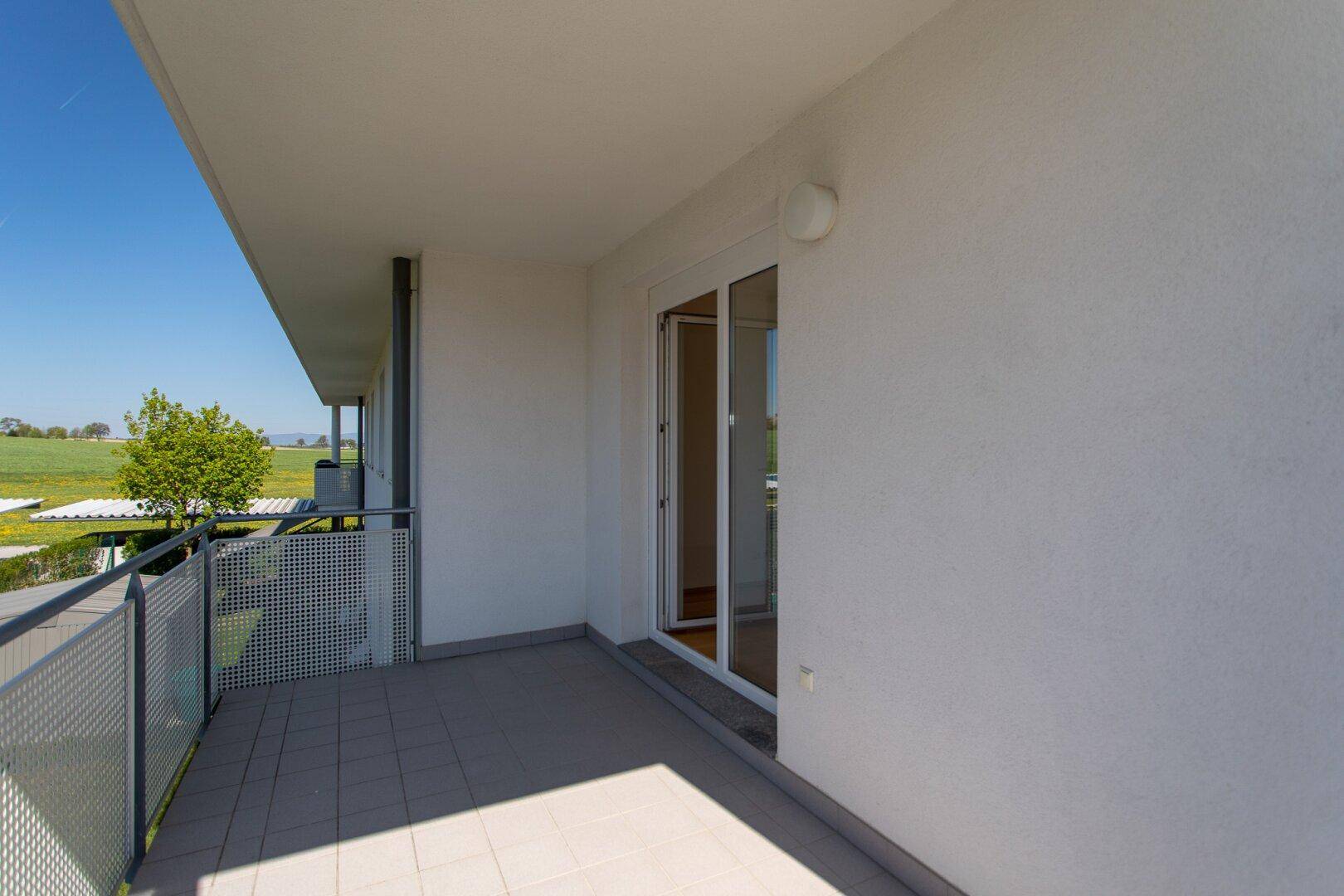Schmaler Balkon mit Fliesenboden und Blick auf grüne Felder unter blauem Himmel.