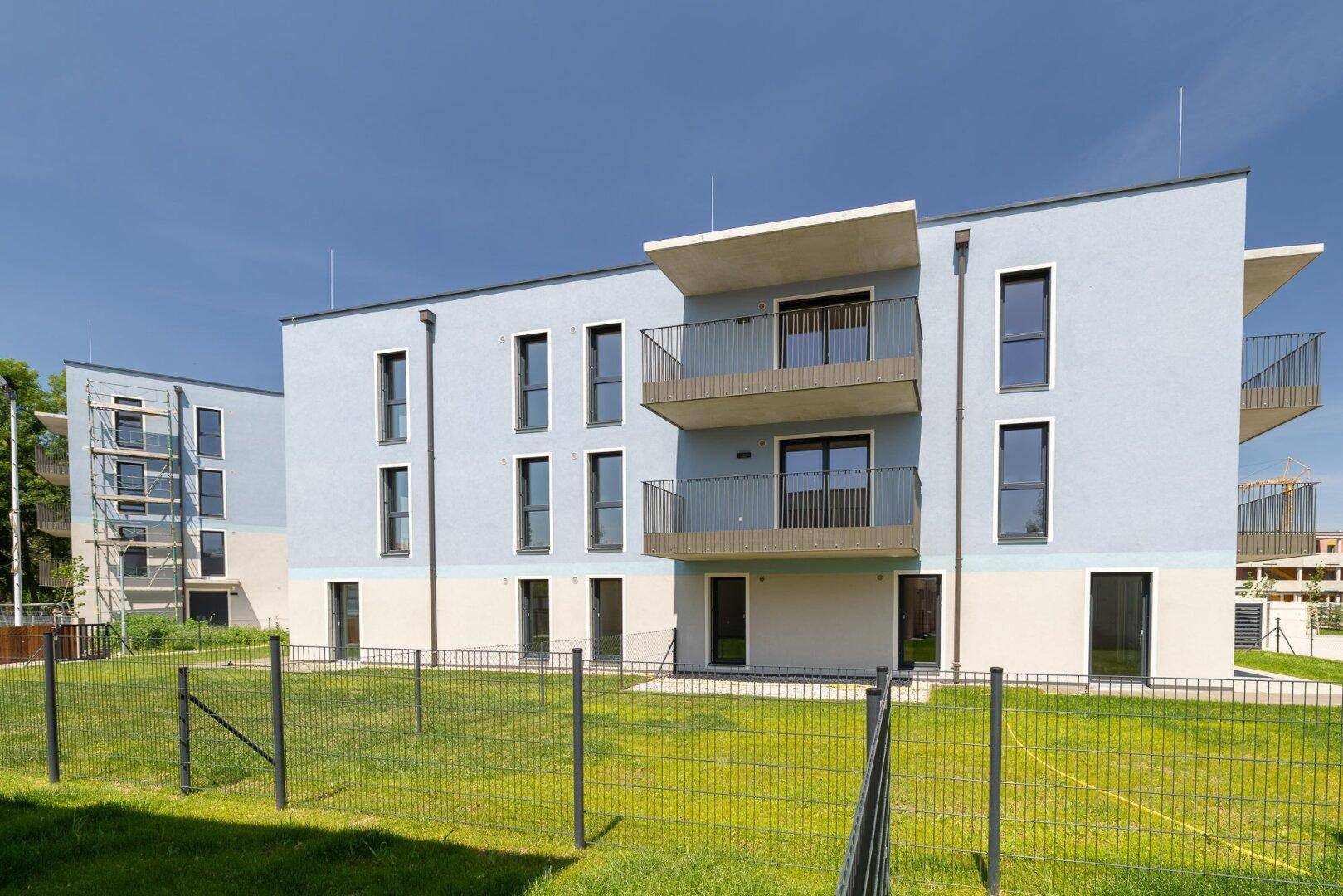 Modernes Mehrfamilienhaus mit hellblauer Fassade, Balkonen und gepflegten Grünflächen unter blauem Himmel.