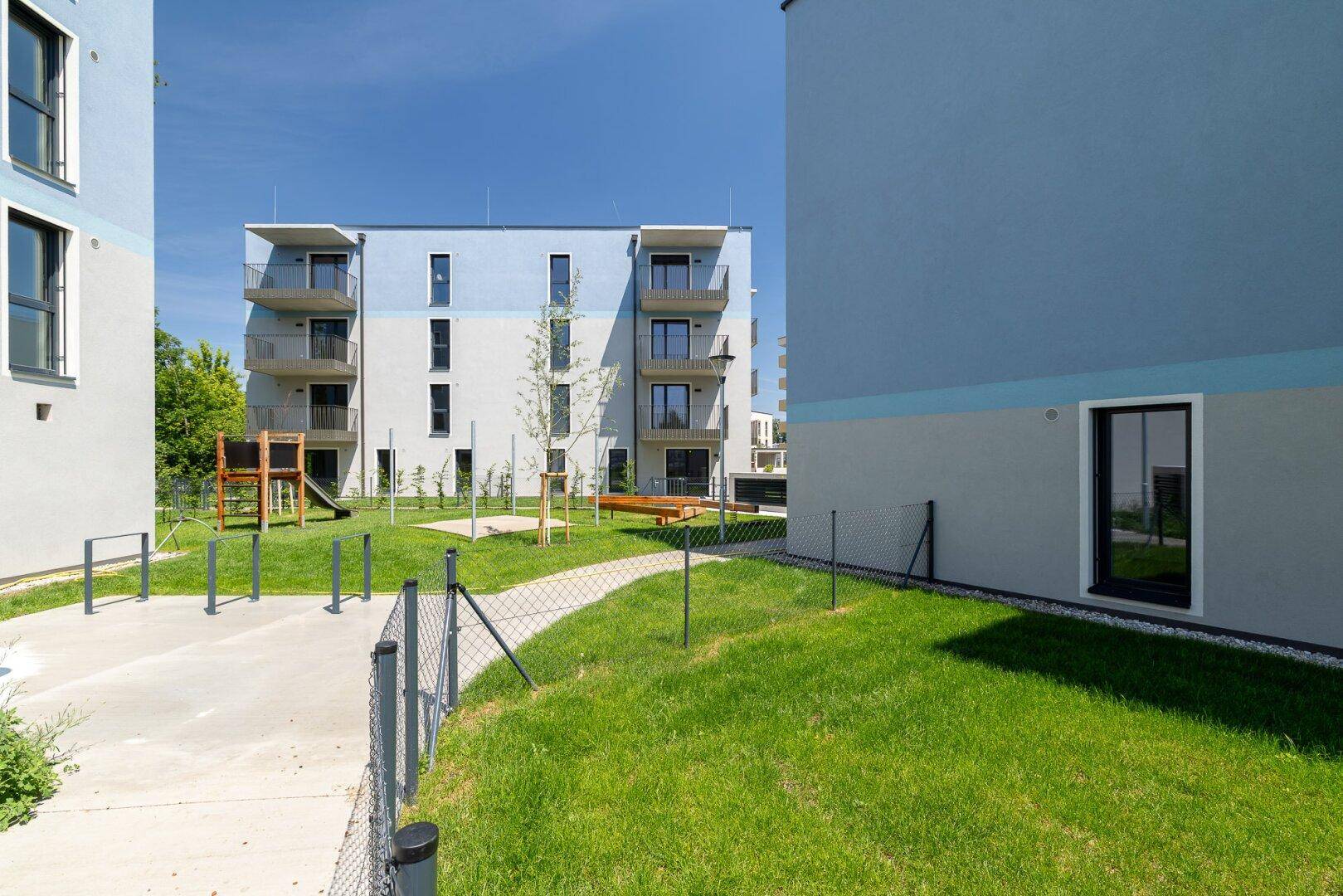 Moderne Wohngebäude mit Spielplatz und Grünflächen in einer neu errichteten Wohnanlage unter blauem Himmel.
