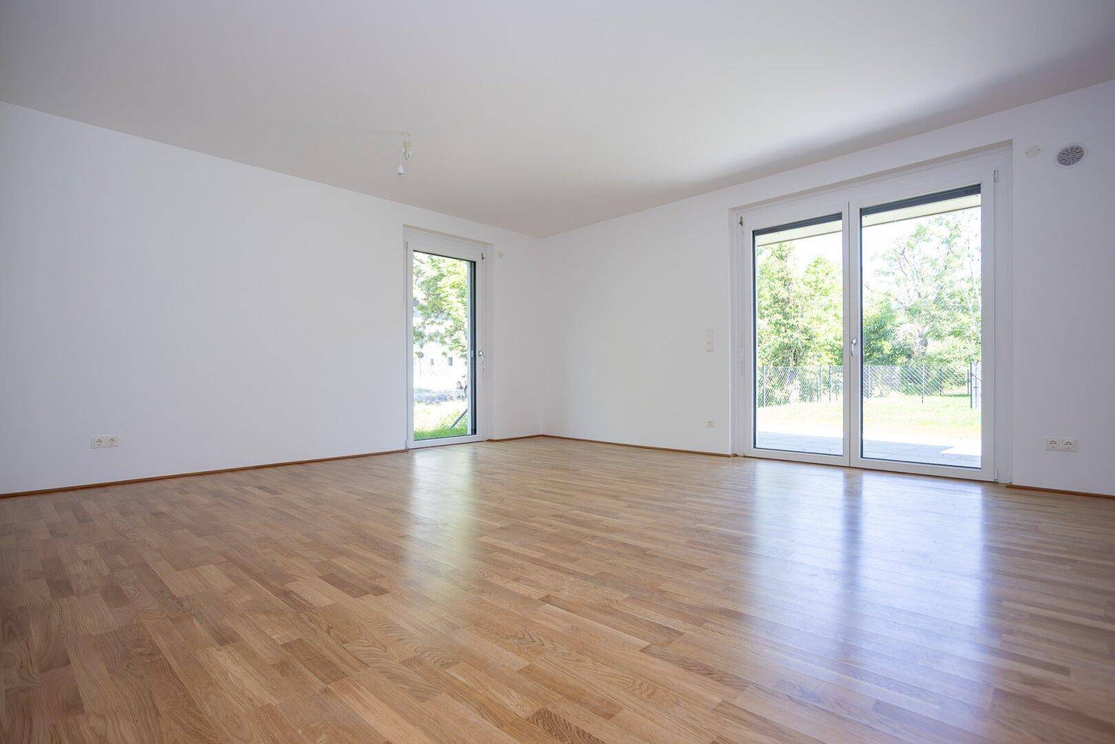Helles, leeres Wohnzimmer mit großen Fenstern und Holzboden, bereit zur individuellen Gestaltung.