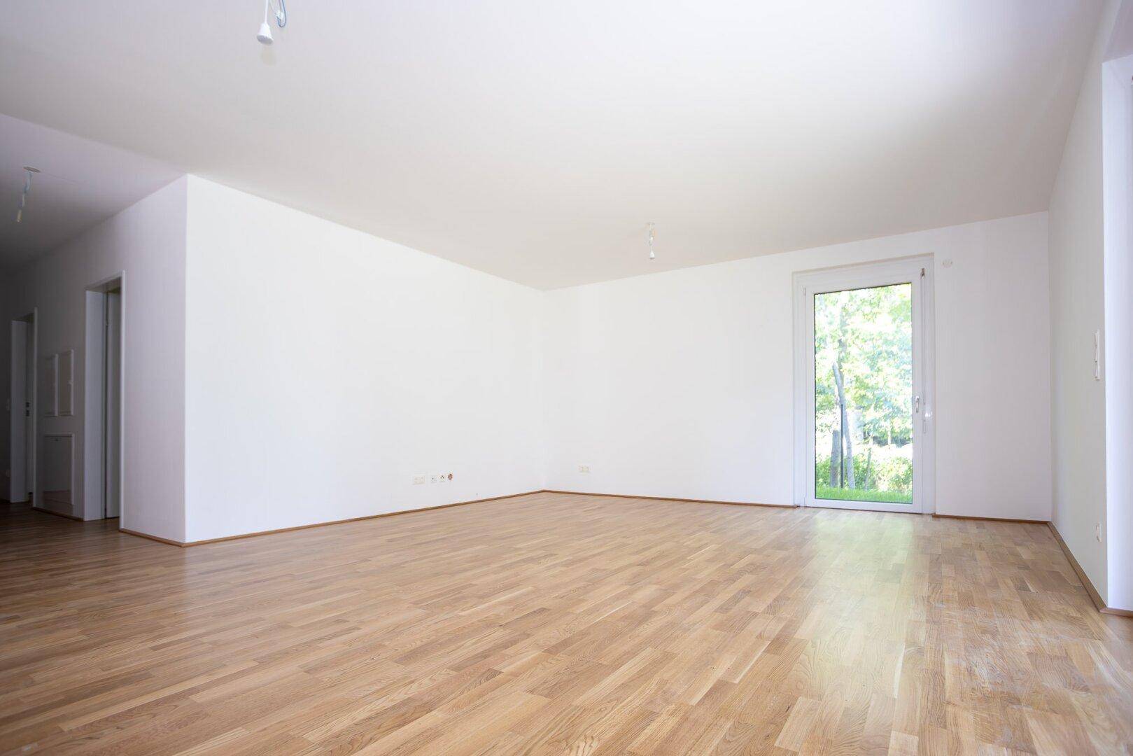 Geräumiges, unmöbliertes Wohnzimmer mit hellem Parkettboden und Zugang zum Außenbereich.