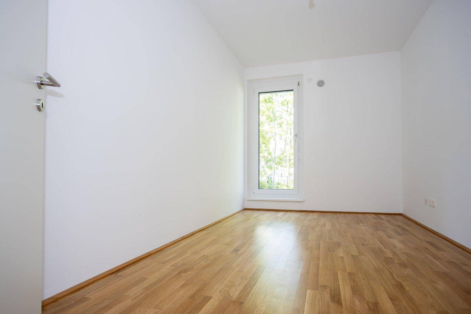 Ruhiges Schlafzimmer mit hellem Parkettboden und großem Fenster, das viel Licht hereinlässt.