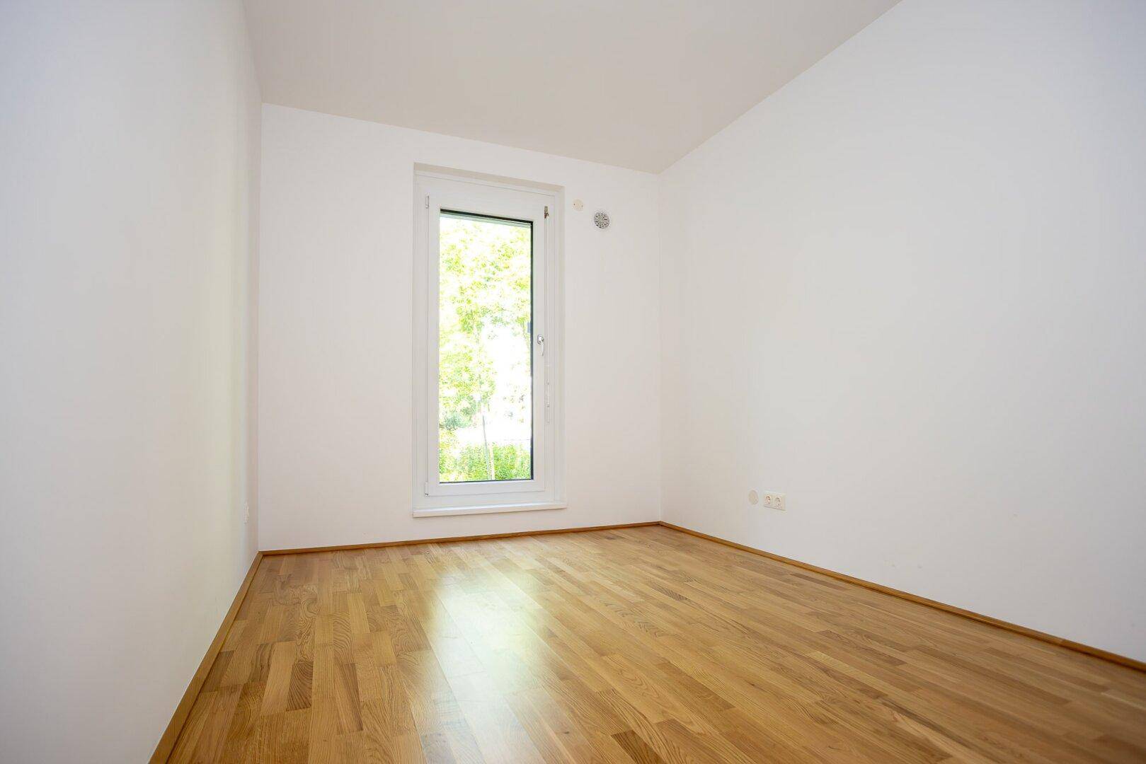 Helles Zimmer mit Parkettboden und Fenster, das einen Blick ins Grüne bietet.