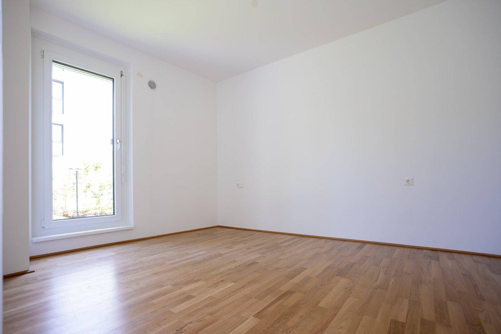 Lichtdurchflutetes Zimmer mit Holzboden und Blick auf die moderne Wohnanlage.