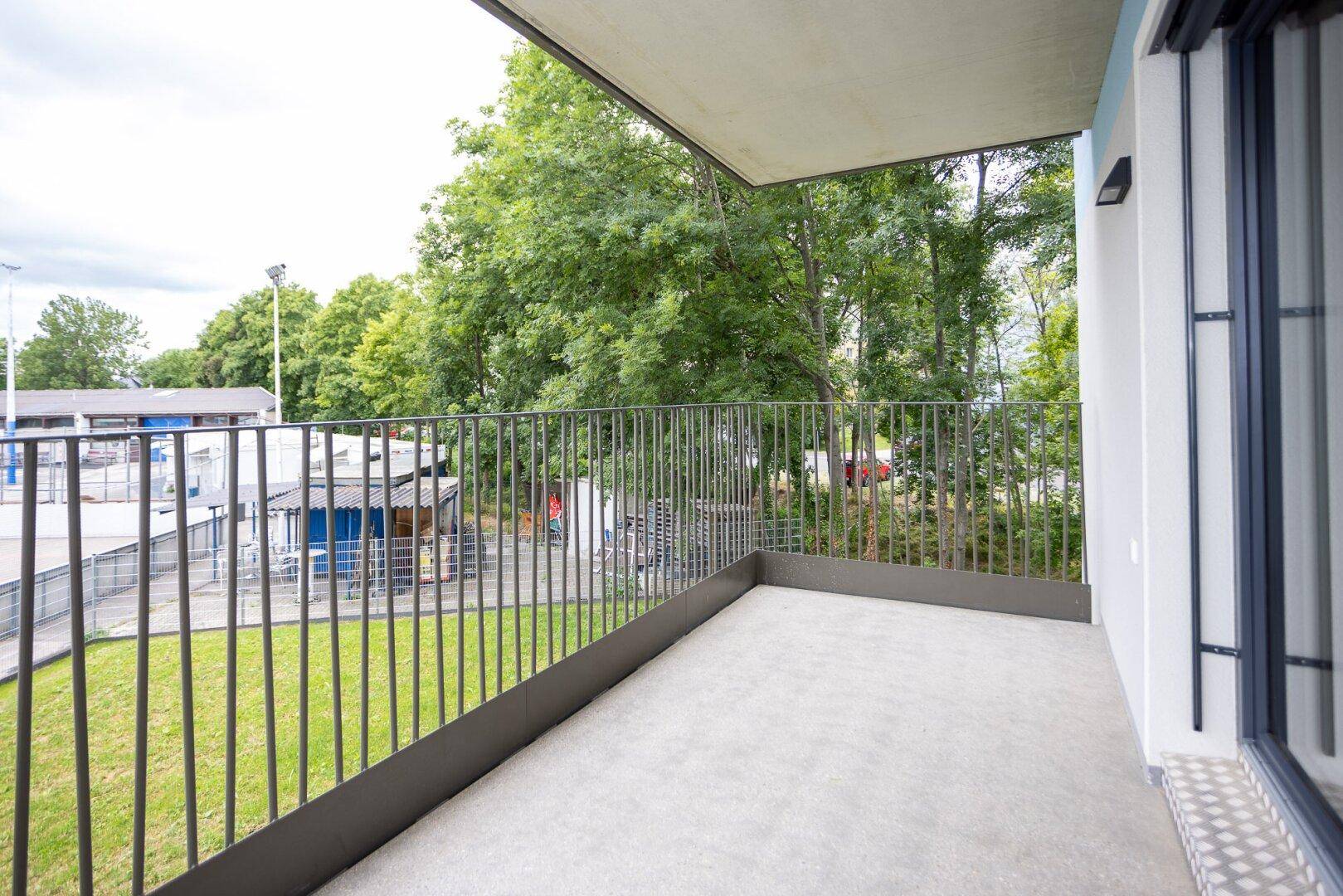 Moderner Balkon mit Metallgeländer und Blick auf grüne Bäume und Sportanlagen.