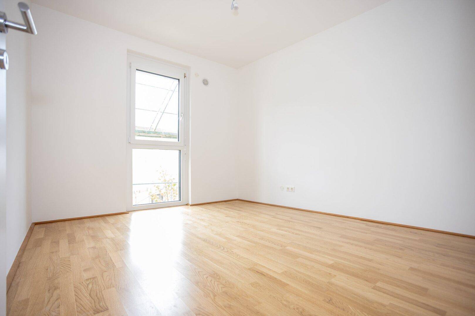 Helles Schlafzimmer mit Parkettboden und einem großen Fenster, bereit zur individuellen Gestaltung.